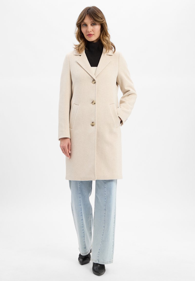 Donna in piedi con un cappotto beige abbottonato sopra un dolcevita nero, jeans blu chiaro e scarpe nere a punta su uno sfondo bianco.