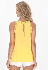 Top giallo senza maniche con bordo in pizzo, dettaglio a chiave sulla schiena e tessuto fluido. Abbinato a pantaloni corti bianchi sfrangiati, che mostrano una trama liscia.