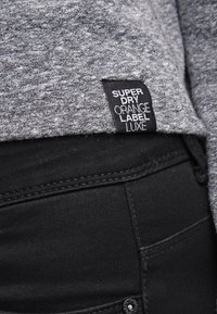 Harmaa neulepaita, jossa on musta etiketti, johon on kirjattu "SUPERDRY ORANGE LABEL LUXE", yhdistettynä mustiin farkkuihin, jotka näyttävät ompelusaumalta ja nappiyksityiskohdilta.