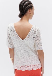 Top bianco in pizzo con maniche corte e schiena a V. Presenta motivi floreali, orlo scallop e tessuto testurizzato. Indossato con pantaloni arancioni.