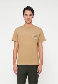 Mees, kes seisab valge tausta ees, kannab beeži graafilise trükiga T-särki ja tumedaid rohelisi pükse, vaatega otse ette ning käed on lõdvestunud.