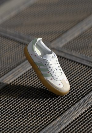 Baskets blanches Adidas Samba avec des rayures vert clair, semelle en gomme, et petits motifs de fleurs rouges sur une grille métallique.