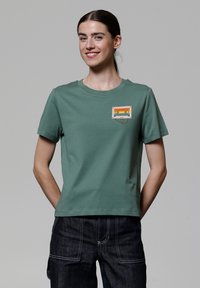 T-shirt en coton vert avec un col rond et des manches courtes. Présente un graphique coloré de cassette audio sur la poitrine gauche.