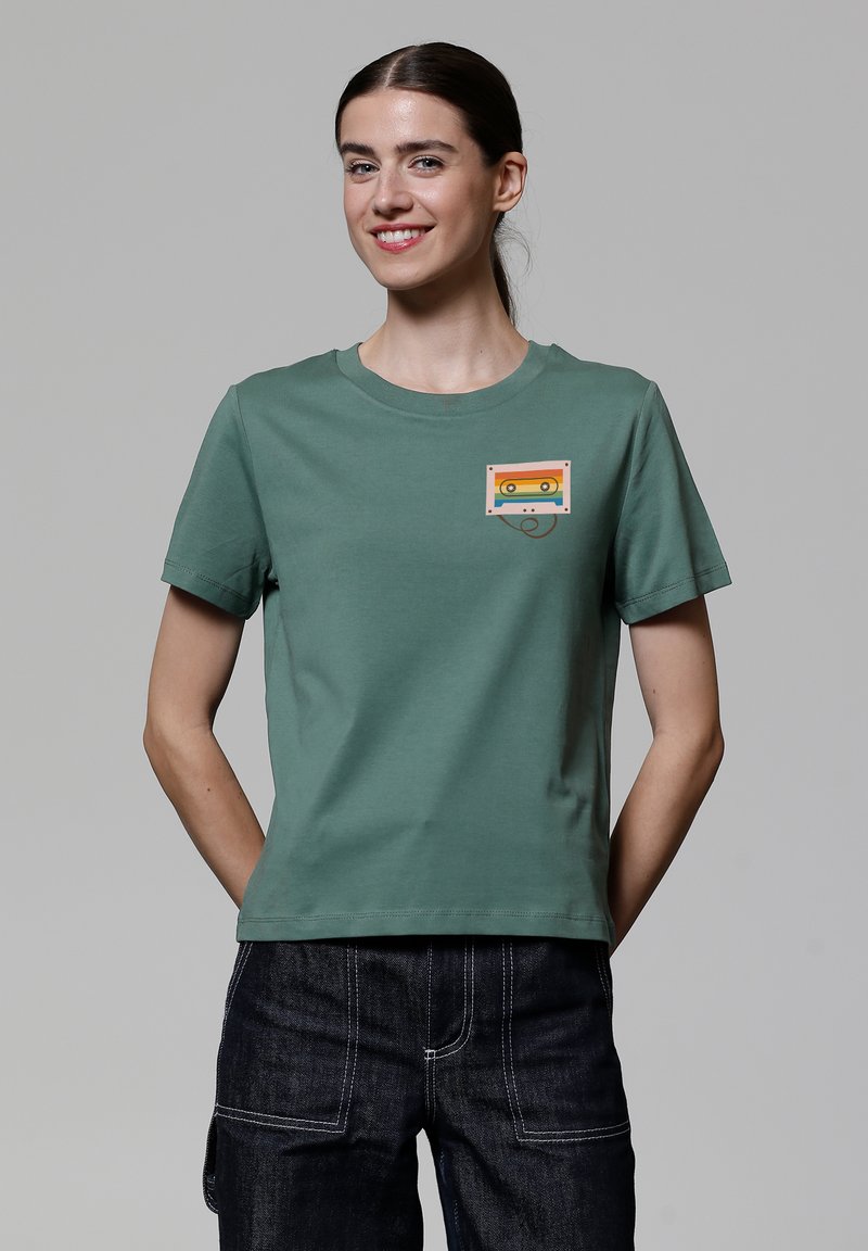 T-shirt en coton vert avec un col rond et des manches courtes. Présente un graphique coloré de cassette audio sur la poitrine gauche.