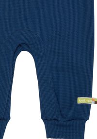 Jumpsuit in tessuto a coste blu navy, con scollo rotondo e maniche lunghe con polsini alle caviglie. Include un piccolo etichetta verde e gialla.