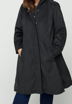 Kobieta nosząca ciemnoszary wełniany płaszcz do kolan z kapturem i rękami w bocznych kieszeniach, w połączeniu z ciemnoniebieskimi dżinsami.