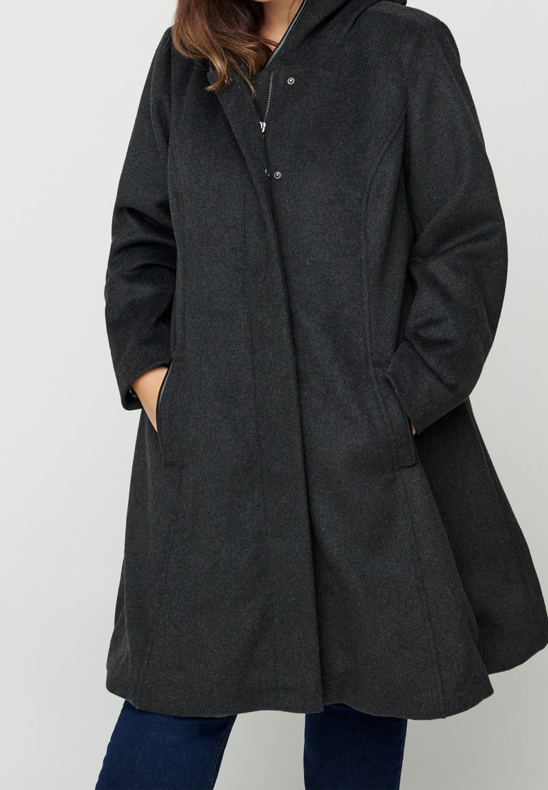 Femme portant un manteau en laine gris foncé longueur genou avec une capuche et les mains dans les poches latérales, assorti à un jean bleu foncé.
