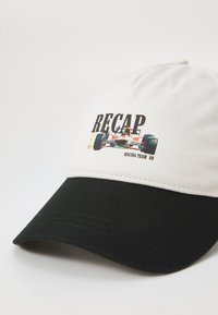 Cappellino con parte superiore bianca e visiera nera. Presenta un design da corsa stampato con il testo "RECAP RACING TEAM '89." Realizzato in tessuto con una texture liscia.