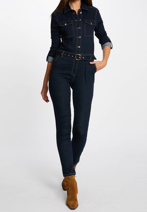 Combinaison en denim foncé à manches longues, avec un devant à boutons, des poches sur la poitrine et une taille ceinturée ; présente des coutures dorées et des poignets roulés bleus.