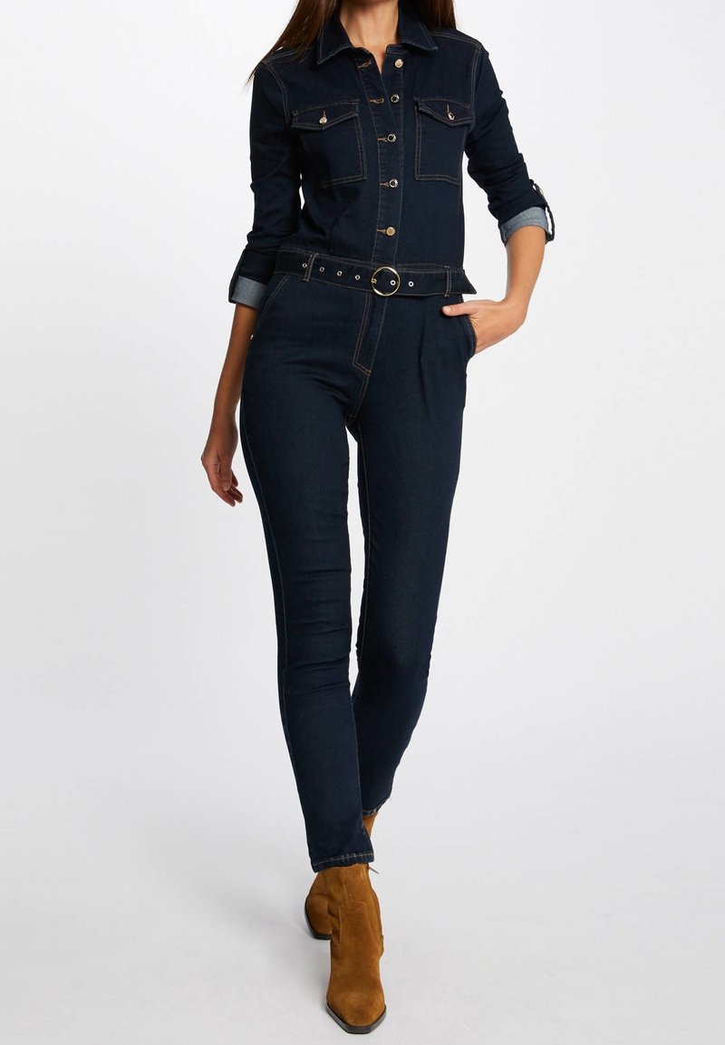 Combinaison en denim foncé à manches longues, avec un devant à boutons, des poches sur la poitrine et une taille ceinturée ; présente des coutures dorées et des poignets roulés bleus.