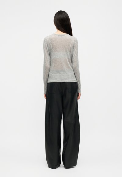 Žena s dlhými tmavými vlasmi, oblečená v svetlosivom tričku s dlhým rukávom a voľných čiernych širokých nohaviciach, stojaca chrbtom k pozorovateľovi na jednoduchom pozadí.