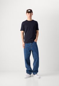 Tricou din bumbac bleumarin, croială lejeră; blugi evazați de un albastru deschis, talie înaltă; adidași albi; șapcă maronie cu logo pe față.