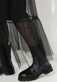 Schwarze Ankle Boots aus strukturiertem Leder, mit robusten Sohlen und Einsätzen aus dehnbarem Stoff. Überlagert von einem durchsichtigen schwarzen Tüll-Stoff.