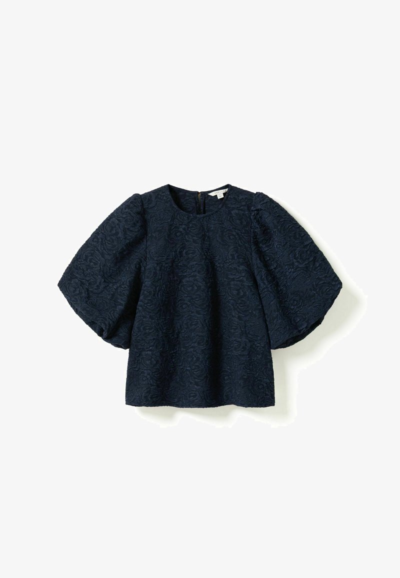 Blouse texturée bleu marine avec col rond et manches courtes bouffantes et volumineuses, ornée d’un motif floral en relief, présentée sur un fond uni.