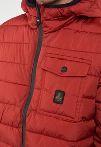 Giacca a vento imbottita rossa con trapuntatura, zip frontale, cappuccio e tasca sul petto con una patta nera e etichetta con logo. Tessuto morbido e lucido.