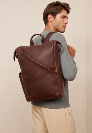 Mann trägt grauen Pullover und beigefarbene Hose und einen großen, dunkelbraunen Leder-Rucksack mit eckigem Design auf dem Rücken.