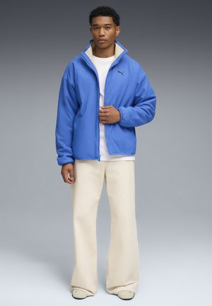Giacca blu con colletto alto e fodera interna morbida, abbinata a pantaloni a gamba larga color crema. Logo PUMA in nero sul petto.