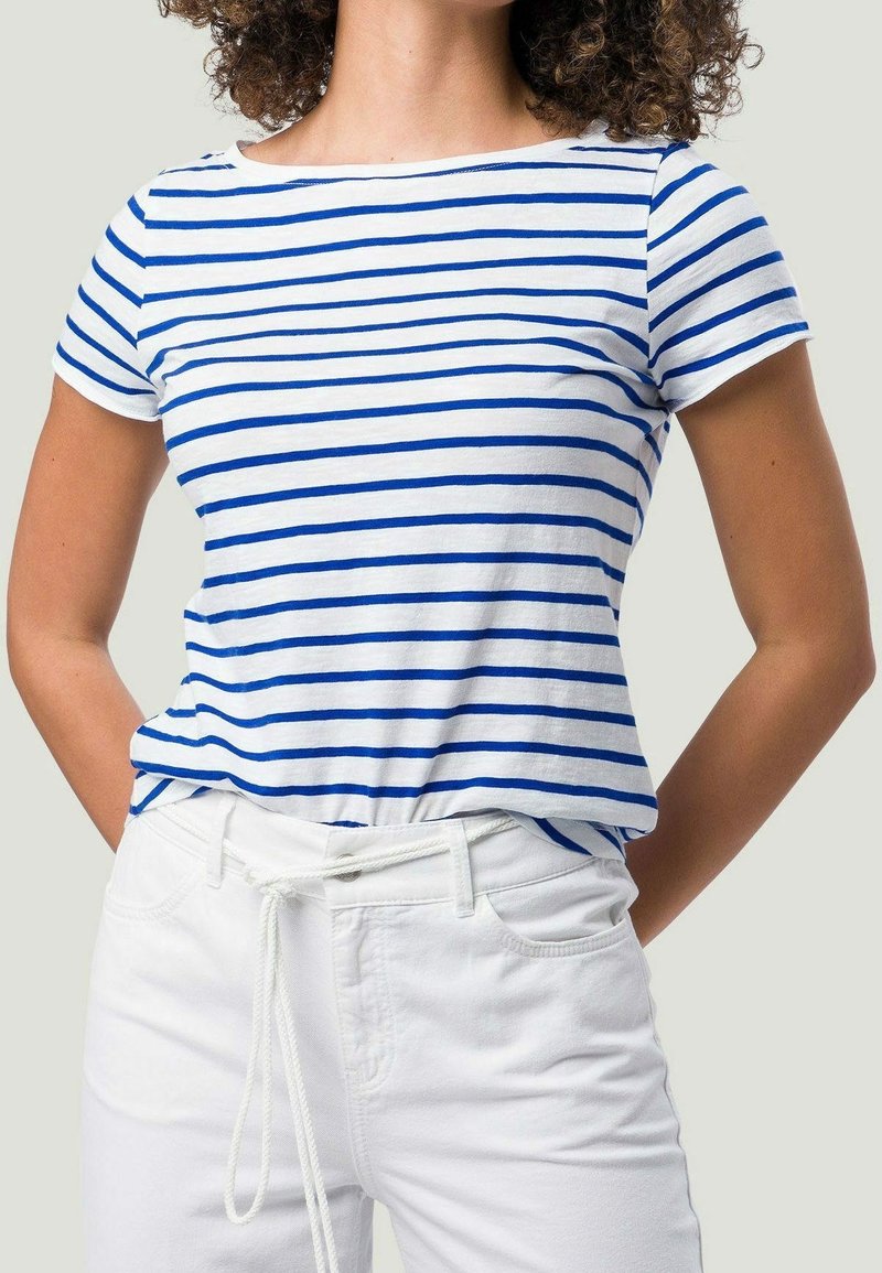 Femme portant une chemise blanche à manches courtes avec des rayures horizontales bleues et un pantalon blanc noué avec une ceinture en corde blanche, les mains derrière le dos.