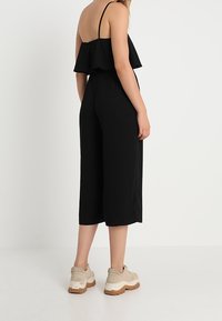 Czarny kombinezon z falbaniastego materiału na górze, szerokimi nogawkami i cienkimi ramiączkami. Noszony z jasnymi trampkami o teksturowanej podeszwie.