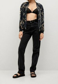 Chaqueta bomber negra con estampado de cadenas doradas, bikini negro, jeans negros de tiro alto y sandalias planas negras. Conjunto casual.