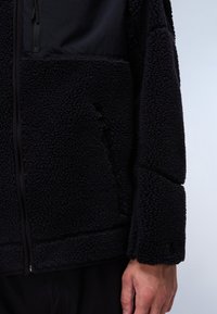 Schwarze Fleecejacke mit einer weichen, strukturierten Oberfläche, ausgestattet mit einer Reißverschlusstasche und kontrastierendem Stoff an der oberen Vorderseite. Nahtdetails sind sichtbar.