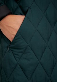 Chaqueta acolchada verde oscuro con un patrón de rombos, que cuenta con un bolsillo con cremallera y una textura suave. Detalles negros en los puños.