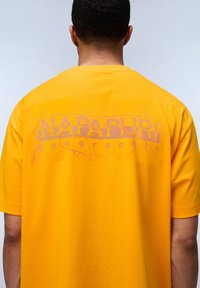 Žluté bavlněné tričko s texturovanou zadní grafikou a nápisem "NAPAPIURI" v kontrastní fialové barvě a volným střihem.