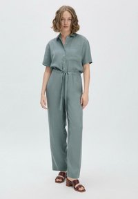 Mintgrüner Jumpsuit mit kurzen Ärmeln, Knopfleiste, Bindegürtel und weitem Bein; kombiniert mit braunen Sandalen mit Absatz. Glatte Stofftextur.