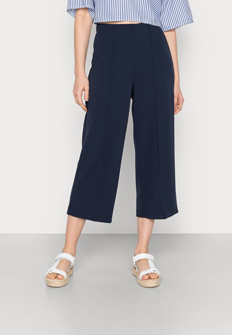 Femme portant un pantalon large court bleu marine, des sandales à plateforme blanches et un top court à rayures bleues et blanches, debout devant un fond uni.