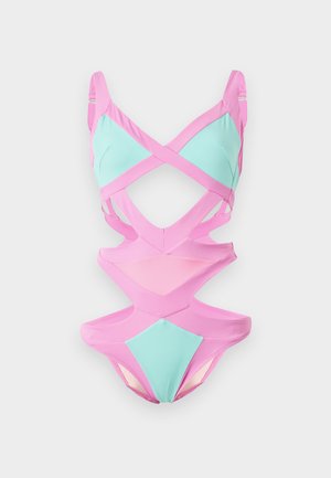 Nő rózsaszín és világoskék kivágott egyrészes fürdőruhában, magas sarkú cipőben, egyszínű fehér háttér előtt áll.