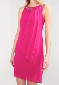 Ärmlös klänning i stark rosa med pärlbesatt halsringning och asymmetrisk framdrapering, bärs av en kvinna med ljus hy och blont hår.