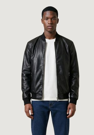 Mladý muž oblečený v černé kožené bomber bundě přes bílé tričko a modré džíny, stojící před jednobarevným světlým pozadím.