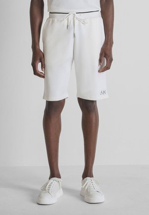 Uomo che indossa pantaloncini bianchi con coulisse e sneakers bianche, in piedi contro uno sfondo chiaro e uniforme.