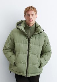 Veste rembourrée en olive verde mute, dotée d'une fermeture éclair à l'avant, d'une capuche ajustable et d'un col côtelé. Conçue en matériau doux et matelassé.