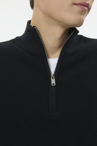 Pulover negru tricotat cu guler cu fermoar pe jumătate, având un material texturat și un fermoar argintiu, purtat peste o cămașă albă.