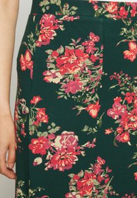 Saia floral em tecido verde escuro com flores vibrantes em rosa, vermelho e creme. Inclui bolsos laterais e uma textura suave.