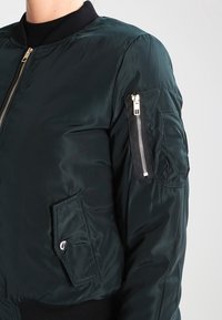 Mörkgrön bomberjacka med dragkedja fram, ribbat krage och flera fickor inklusive en ficka med dragkedja på ärmen. Slät, glänsande tyg.