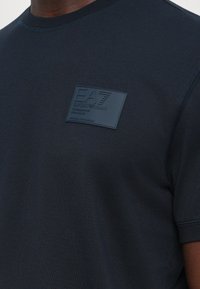 Detailní záběr na tmavě modré tričko s obdélníkovou nášivkou loga EA Emporio Armani na hrudi.