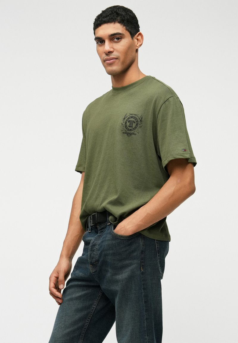 Olivgrön kortärmad t-shirt med en svart grafiklogotyp på bröstet, tillsammans med mörkblå denimjeans och ett svart bälte.