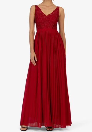 Kobieta w czerwonej, długiej sukni bez rękawów z koronkowym kwiatowym gorsetem i plisowaną spódnicą, w czarnych sandałach na szpilce z paskami.
