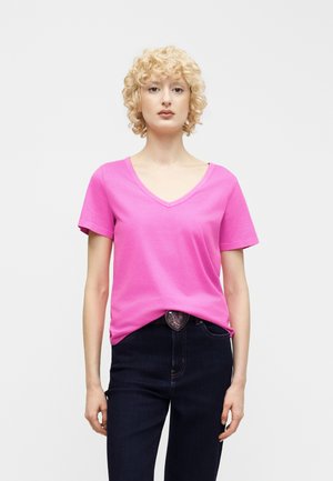 Osoba o blond kręconych włosach, ubrana w jasnoróżowy T-shirt z dekoltem w serek, lekko wpuszczony w ciemne dżinsy, z klamrą paska w kształcie serca.