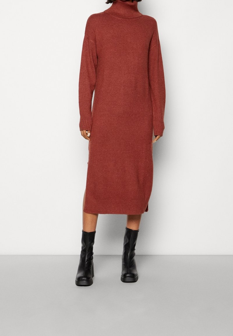 Robe pull à col roulé en maille rouge brique, dotée de manches longues, de poignets côtelés et d'une fente sur le côté. Associée à des bottes noires montantes jusqu'au genou.