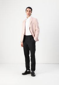 Jasnoróżowy blazer, biały shirt oraz czarne, dopasowane spodnie. Czarne buty uzupełniają strój. Model stoi na tle jednolitego tła.
