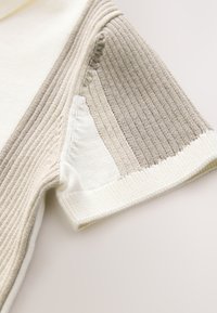 Adīta džempera aproce baltās un bēšās toņos ar reljefām ribām un horizontālām svītrām. Mīksts audums ar dažādu adījumu rakstiem.