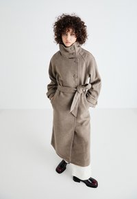 Casaco longo de lã castanho, com gola alta, dois botões, cintura cingida e bolsos laterais; modelo usa sapatos pretos e calças brancas.