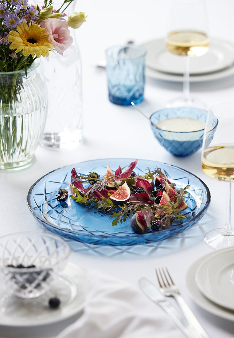 Niebieski szklany talerz z teksturowanym brzegiem, zawierający sałatkę z figami, jagodami i zielenią. W otoczeniu eleganckie naczynia i kwiaty.