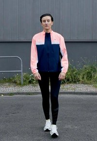 Chaqueta atlética de color navy y rosa con cremallera, combinada con leggings negros y zapatillas de correr blancas. Tela suave, diseño ajustado, marca mínima.