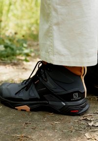 Scarpa da trekking nera con design alto, caratterizzata da tessuto testurizzato, accenti arancioni a contrasto e suola robusta. Adatta per l'uso all'aperto.