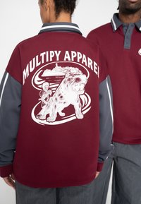Dunkelrotes Langarmshirt mit grauen Akzenten zeigt eine weiße Grafik eines Hundes und einer Figur, mit dem deutlich sichtbaren Text "MULTIPY APPAREL".