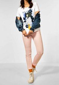 Bílý grafický tričko s barevným potiskem, džínsová bunda, světle růžové kalhoty a béžové tenisky se šňůrkami. Viditelné oranžové ponožky.